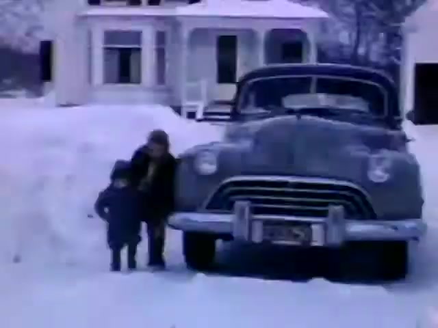 The still from 'Connie & Patty #2' features a snowy scene with a woman and a child near a vintage car. The architecture in the background suggests a rural setting, reminiscent of mid-20th century homes. The car's design, likely from the late 1940s, supports a dating of the film to the post-World War II era. The overall mood and clothing hint at a familial, winter outing during that time period.