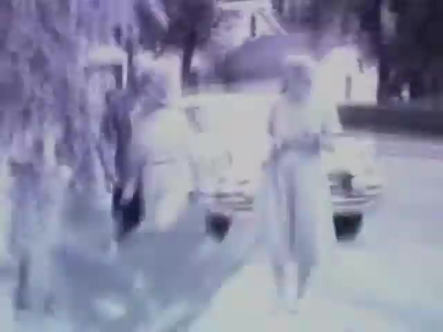 The still from 'Pat in Ocean - 1955' depicts a black-and-white scene where a group of individuals is seen near a parked car on a street. The figures appear to be engaging with one another, possibly in conversation. The surrounding environment features greenery and residential buildings, contributing to a nostalgic 1950s atmosphere. The image has a slightly blurred appearance, enhancing its vintage aesthetic.