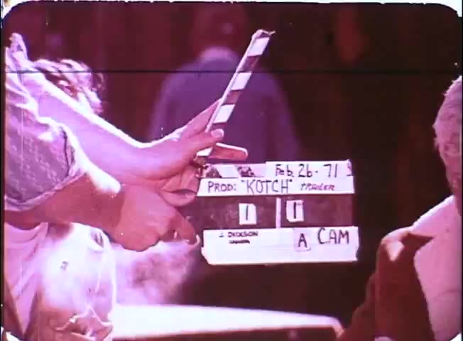 The still depicts a film set scene from the promotional material for 'Kotch,' featuring a clapperboard that reads 'Feb. 26, 71,' indicating the production date. The clapperboard is prominently held up, with 'Prod: KOTCH' and 'A CAM' written on it. In the background, indistinct figures are visible, creating a candid atmosphere typical of behind-the-scenes film shoots. The color palette suggests a vintage 16mm film aesthetic.