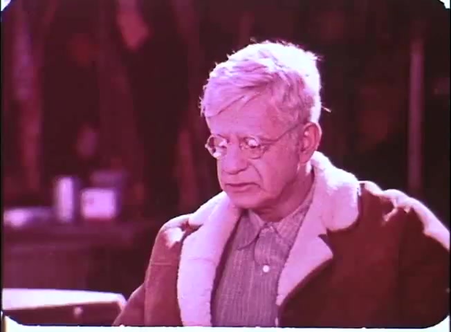 The still features a character depicted with gray hair, wearing glasses and a shearling coat. The expression on his face suggests contemplation or concern, set against a blurred background that hints at an outdoor scene. The color palette is washed out, typical of a 16mm print, giving the image a nostalgic quality.