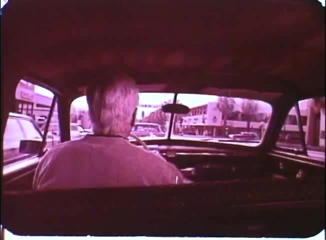 The still shows a view from inside a car, focusing on an older man driving, with his back to the camera. The scene captures a street bustling with cars and buildings in the background. The color tone has a vintage, reddish hue typical of 16mm prints, evoking a nostalgic feel consistent with the promotional material for 'Kotch' featuring Walter Matthau.
