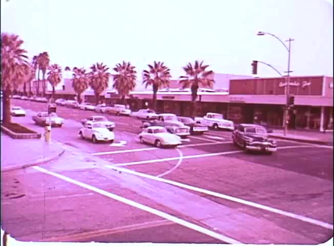The still from the 16mm print of 'Kotch' features a sunny street scene with palm trees lining the road. Multiple vintage cars are seen driving along a bustling intersection, highlighting a mid-20th century urban setting. The overall tone is bright and nostalgic, indicative of the film's time period. Shops with signage can be seen in the background, contributing to the lively atmosphere.