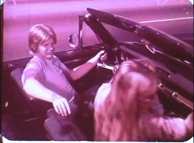 The still features a young man driving a convertible car, with a young woman leaning towards him. The scene has a pinkish hue, typical of the 16mm print aesthetic. The background suggests a road setting, giving a sense of movement. The characters appear engaged with each other, hinting at a casual or intimate moment.