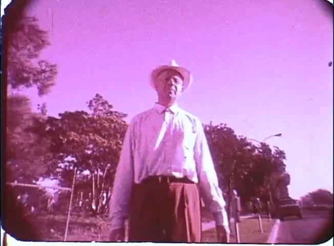 The still from 'Kotch' features a man, likely Walter Matthau, standing outdoors in bright daylight. He is dressed in a light shirt and a wide-brimmed hat, exuding a relaxed yet contemplative demeanor. The image has a pinkish tint, likely due to the 16mm print's age or color grading, and in the background, there are trees and a few parked cars, suggesting a suburban setting. The overall composition captures a moment of solitude or introspection.
