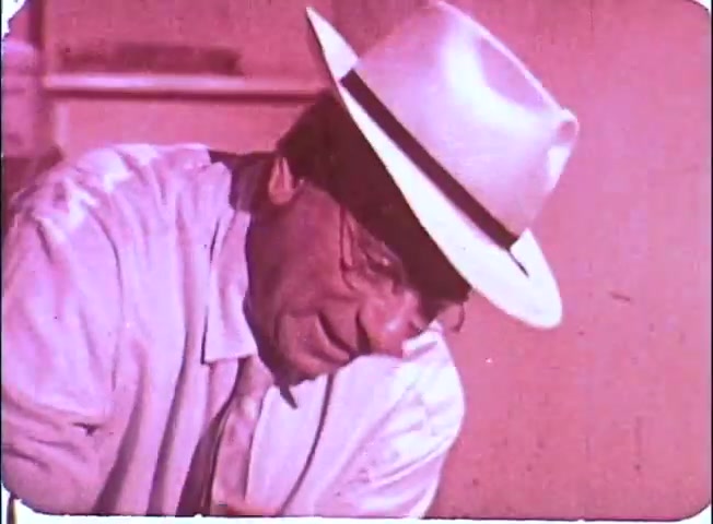 The still features a middle-aged man wearing a light-colored fedora and a white shirt, leaning slightly forward. His expression appears thoughtful or focused, conveying a sense of contemplation. The image exhibits a warm, muted color palette typical of 16mm film, adding a nostalgic feel to the scene. There’s a soft focus, contributing to the vintage aesthetic characteristic of promotional materials from the era.