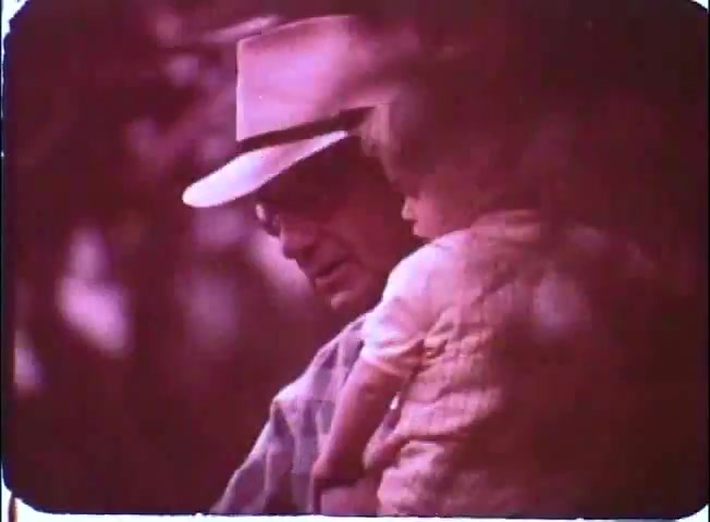 The still from 'Kotch' features a man, likely depicted as Walter Matthau, wearing a light-colored hat and glasses, holding a young child. The scene has a warm, nostalgic tone, with soft colors and a blurred background, suggesting a tender moment between the characters amidst a serene setting. The focus on their interaction conveys warmth and connection.