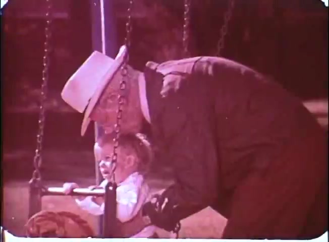 The still from 'Kotch' features an older man with glasses and a white hat leaning close to a young child seated in a swing. The child, appearing joyful, is focused on the activity. The vibrant colors and soft focus suggest a nostalgic or heartwarming atmosphere, emphasizing the gentle interaction between the two in a park setting. The scene encapsulates themes of innocence and connection across generations.