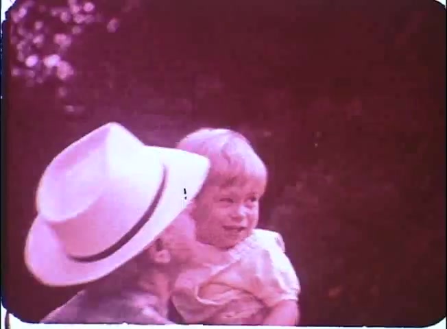 The still features a man wearing a white hat affectionately kissing a young child, who is smiling joyfully. The scene has a nostalgic, warm tone typical of 16mm prints, with soft colors that suggest a casual and intimate moment between them, likely conveying themes of family and connection from the film 'Kotch.' The background appears blurred, emphasizing the tender interaction.