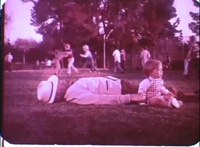 The still captures a park scene with a subtle, nostalgic feel typical of 16mm prints. In the foreground, an older man, presumably Walter Matthau's character, is laid back on the grass, wearing a white hat and light-colored clothing. Nearby, a small child in a checkered outfit sits beside him, seemingly relaxed. The background is lively, filled with people engaging in various activities like playing and socializing, adding a sense of warmth and community to the image. The colors have a slightly faded quality, enhancing the vintage aesthetic of the film.