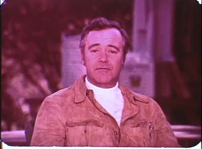 The still features Walter Matthau, seated and facing the camera. He is wearing a light-colored shirt beneath a tan jacket, exuding a casual, approachable demeanor. The background is softly blurred, suggesting an outdoor setting. The image has a vintage quality typical of 16mm prints, with slight color fading, enhancing its nostalgic feel.