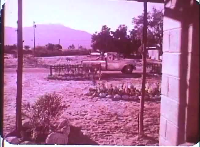 The still from 'Kotch' features a dusty outdoor scene, likely set in a rural area. A vintage pickup truck is seen parked along a road, with a backdrop of mountains in the distance. The foreground showcases a garden with neatly arranged plants and rocks, creating a tranquil, nostalgic atmosphere. The overall color tone carries a warm, faded look typical of a 16mm print.
