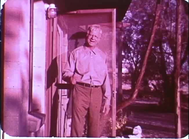 The still depicts a smiling older man standing at the entrance of a house, suggesting a warm and inviting atmosphere. He wears a light-colored shirt and trousers, and there are hints of rustic surroundings with trees and natural elements in the background. The color palette has a vintage quality typical of 16mm film, enhancing its nostalgic feel.