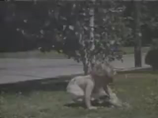 The still depicts a young child playing on grassy ground, possibly engaged with a small animal. The scene is brightened by sunlight filtering through nearby trees, with a subtle backdrop of a street. The vibrant Kodachrome colors capture a nostalgic, carefree moment from 1941.