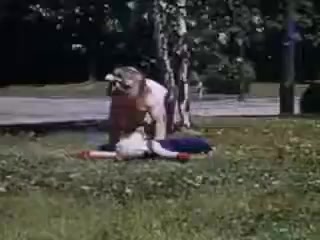 The still from 'Connie and Shirley reel #1 - 1941' features a vibrant, colorful Kodachrome scene set outdoors. It captures two children, one apparently on the ground and the other seemingly standing over or playing with them. The background includes trees and a glimpse of a road, suggesting a leisurely, playful moment typical of childhood in the early 1940s. The colors are rich and the composition suggests a candid, joyful interaction between the children.