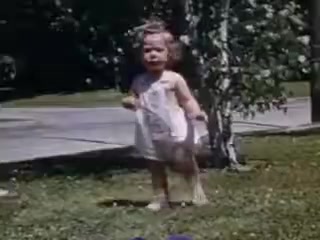 The still from 'Connie and Shirley reel #1 - 1941' depicts a young child, likely a toddler, standing in a grassy area. The child is wearing a light-colored outfit and appears to be playfully engaged, with a slight smile or expression on their face. In the background, there is a small tree and hints of residential elements like a road and possibly a house. The vibrant colors characteristic of Kodachrome film capture a joyful, nostalgic moment from the early 1940s.