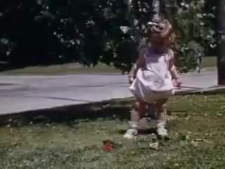 The still features a young girl in a light dress and shoes, playing on grassy ground. She appears to be in a suburban setting, with a road visible in the background. The vibrant Kodachrome colors suggest a sunny day in 1941, capturing a moment of childhood innocence and outdoor play.