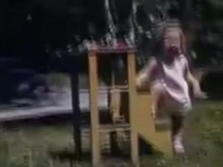 The still shows a young child in a light-colored outfit, seemingly mid-action, possibly playing outdoors. She is jumping or climbing from a yellow play structure. The scene is vibrant with lush green grass and trees in the background, indicative of a carefree, joyful moment in a childhood setting from 1941. The Kodachrome film captures the colors vividly, reflecting the era's playful and innocent ambiance.