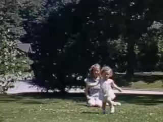 The still from 'Connie and Shirley reel #1 - 1941' captures a nostalgic moment in a sunlit outdoor setting, featuring two young girls playing on a grassy lawn. One girl sits with her legs crossed while the other runs towards her, both dressed in light-colored, playful outfits. The background includes trees and a glimpse of a house, evoking a serene summer day atmosphere typical of the early 1940s. The vibrant colors highlight the cheerful innocence of childhood during that era.