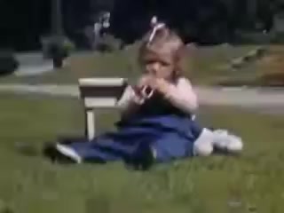 The still depicts a young girl seated on the grass, wearing a light-colored top and a blue skirt. She appears to be playing with something in her hands, possibly a toy. The background features a park-like setting with some blurred figures in the distance, suggesting a leisurely, outdoor scene typical of childhood play in the early 1940s. The image is vibrant, showcasing the warm, saturated colors characteristic of Kodachrome film from that era.