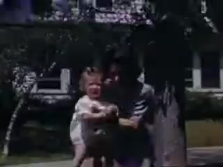 The still captures a tender moment between a woman and a child in a sunny outdoor setting. The woman, likely a caregiver or parent, holds the child, who appears to be smiling with a joyful expression. The background features a well-kept lawn and some trees, suggesting a suburban or family-friendly environment typical of the early 1940s. The colors are vibrant, indicative of Kodachrome film stock.