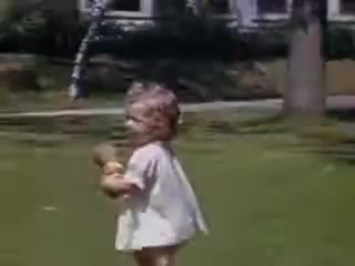 The still features a young child outdoors, smiling as they run on grassy terrain. The child holds a doll and wears a light-colored outfit. In the background, there are trees and a house, suggesting a bright, sunny day in a residential area. The colors are vibrant, characteristic of Kodachrome film from 1941.