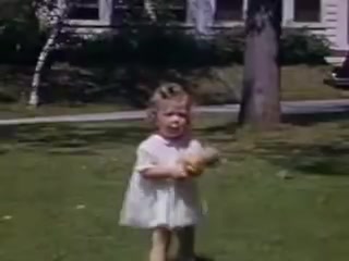 The still features a young girl in a white dress, standing on a grassy lawn. She has curly hair and is holding a toy. In the background, there are trees and a house, suggestive of a suburban setting in 1941, captured in vibrant Kodachrome color. The scene conveys a sense of childhood playfulness and innocence.