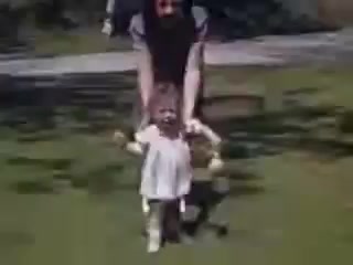 The still shows a joyful moment from the 1941 film 'Connie and Shirley reel #1.' A woman appears to be playfully assisting a toddler, who is wearing a light dress, as they walk together on a grassy area. The scene captures a sense of laughter and childhood innocence, with natural light and vibrant Kodachrome colors enhancing the nostalgic atmosphere of the early 1940s.