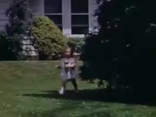 The still from 'Connie and Shirley reel #1 - 1941' features two children playing outdoors on a grassy lawn. The vibrant colors of the Kodachrome film highlight the lush greenery and shrubbery in the background. The setting appears to be a residential area, with a spacious home visible, indicative of the time period. The children seem to be engaged in playful activity, capturing a moment of joy and innocence typical of childhood.