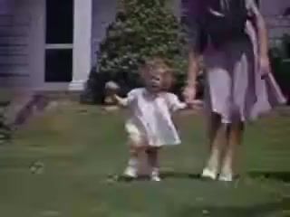 The still from 'Connie and Shirley reel #1 - 1941' depicts a young child in a light dress, walking on a green lawn while holding the hand of an adult. The adult wears a skirt and appears to guide the child. The background features a house and lush foliage, characteristic of a suburban setting in the early 1940s. The image captures a candid moment, showcasing family interaction and a vibrant Kodachrome color palette typical of that era.