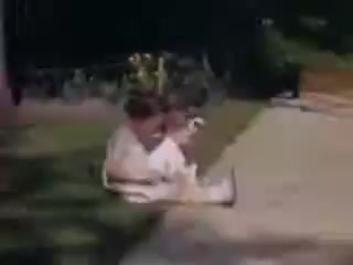 The still captures two children, likely Connie and Shirley, sitting on a grassy area next to a sidewalk. They appear to be engaged in a playful activity, with trees and flowers in the background. The vivid colors typical of Kodachrome film enhance the nostalgic atmosphere of the scene, highlighting a moment of childhood joy from 1941.