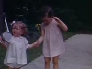 The still from 'Connie and Shirley reel #1 - 1941' features two young girls, dressed in light-colored outfits, walking hand in hand along a pathway. One girl is holding a small object, possibly a toy, while the other appears to be playfully gesturing. The scene captures a moment of childhood innocence, set against a lush, green background typical of outdoor play in the 1940s. The vibrant colors of the Kodachrome film evoke a nostalgic feel.
