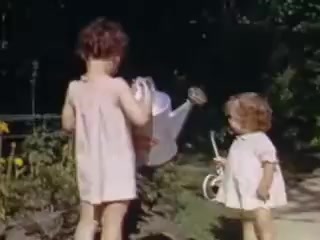 The still features two young girls in a garden setting, embodying a nostalgic scene from 1941. The older girl is holding a metal watering can, while the younger girl stands nearby, clutching a toy. Both are dressed in light-colored, simple clothing, reflecting the casual style of the era. The garden is lush with greenery and flowers, creating a vibrant and cheerful atmosphere. The colors are warm and vivid, characteristic of Kodachrome film.