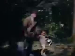 The still from 'Connie and Shirley reel #1 - 1941' depicts two figures, likely a child and an adult, amidst a natural setting. The scene is captured in vibrant Kodachrome colors, typical of the 1940s, showcasing the lush greenery surrounding them. The adult appears to be holding the child, conveying a sense of playfulness and intimacy in this candid moment. The lighting suggests it might be a sunny day, enhancing the nostalgic quality of the image.