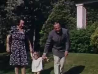 The still shows a cheerful scene from 1941, featuring an adult woman and man holding hands with a young child in a lush, green lawn. The woman is wearing a patterned dress, while the man is in a suit. The child, dressed in a light outfit, appears joyful as they walk together. In the background, there are trees and a glimpse of a house, typical of a suburban setting from that era. The colors are vibrant, characteristic of Kodachrome film.