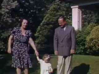 The 16mm still from 'Connie and Shirley reel #1 - 1941' features a family scene outdoors. A woman in a patterned dress is holding hands with a small child, who is wearing a light-colored outfit. Beside them stands a man in a suit. The setting appears to be a well-maintained garden, with lush greenery in the background, suggesting a joyful family moment captured in vibrant Kodachrome colors from the early 1940s.