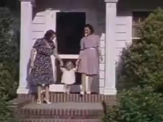 The still from 'Connie and Shirley reel #1 - 1941' features two women standing on the steps of a house, accompanied by a small child. The women are dressed in period-appropriate attire: one in a floral dress and the other in a pastel dress with a belt. The child, wearing a white outfit, is grasping the hand of one woman as they appear to step out together, suggesting a casual family moment. The setting includes a simple porch with white columns and greenery in the foreground, highlighting a typical suburban environment of the era. The vibrant Kodachrome colors enhance the nostalgic feel of the scene.