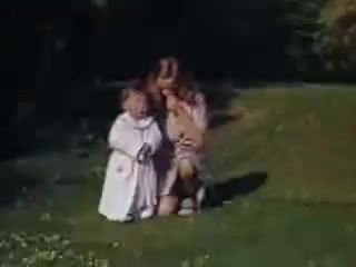 The still from 'Connie and Shirley reel #1 - 1941' features two young girls sitting on green grass. One girl is dressed in a white outfit, while the other wears a light-colored dress. They appear to be engaged in a playful or intimate moment, surrounded by lush greenery, typical of outdoor scenes from that era. The vibrant colors suggest the use of Kodachrome film, highlighting the joy of childhood in a picturesque setting.