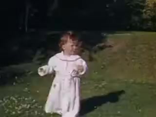 The still from 'Connie and Shirley reel #1 - 1941' features a young child dressed in a white outfit, happily running across a grassy area. The scene is bathed in warm, natural light, typical of Kodachrome film, showcasing vibrant colors. The background includes hints of greenery and a gently sloping hill, creating a cheerful, idyllic outdoor setting.