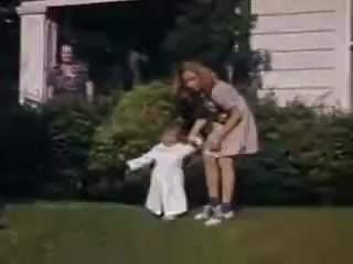 The still from 'Connie and Shirley reel #1 - 1941' features a young girl helping a toddler walk in a lush green yard. The toddler is dressed in a white outfit, while the older girl wears a simple dress and sneakers. In the background, two adults can be seen observing, adding a familial atmosphere to the scene. The vibrant colors of Kodachrome highlight the joyful and lively nature of this moment.