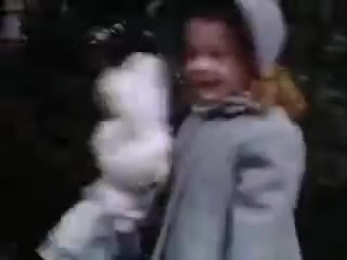 The still from 'Connie and Shirley reel #1 - 1941' features a young girl dressed in a light blue coat and a hat, holding a doll. The image captures a moment of joy, with the girl smiling against a backdrop that suggests outdoor scenery. The use of Kodachrome film adds a vintage, colorful quality to the scene, characteristic of early 1940s home movies.