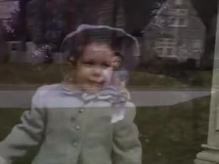 The still features a young child at play, dressed in a light-colored outfit with a hat, smiling and engaging with the camera. There is a blurred background that suggests a lush garden or park setting, typical of early 20th-century family outings. The colors are vibrant, characteristic of Kodachrome film from 1941, giving it a nostalgic and warm feel.