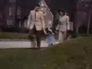 The still captures a family strolling along a path in a suburban setting from 1941. A man in a light suit and a woman in a light dress walk hand-in-hand with a young girl in a blue dress. The background features a house with architectural details typical of the era, surrounded by greenery. The colors are vibrant, characteristic of Kodachrome film, evoking a nostalgic atmosphere.
