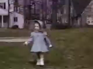 The still captures a young girl in a light blue dress and white shoes, joyfully running on a grassy area. She appears to be in a suburban setting, with trees and a house in the background, reflecting the nostalgic charm of 1941. The Kodachrome film gives the image a vivid, warm color palette typical of the era.