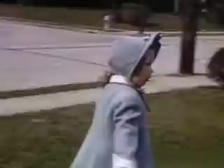 The still shows a child dressed in a light blue coat and matching bonnet, walking on a grassy area near a sidewalk. The setting appears to be suburban, with a road visible in the background. The use of Kodachrome film suggests vibrant colors typical of early 1940s home movies. The overall mood is nostalgic, capturing a moment from childhood in 1941.