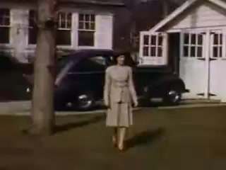 The still captures a woman in a stylish light-colored dress walking on a lawn, set against a suburban backdrop from the early 1940s. A classic car is parked nearby, and there are two houses in the background, indicative of the architecture and lifestyle of that period. The vibrant colors suggest the use of Kodachrome film, reflecting the era's aesthetic.