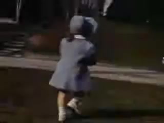 The still captures a young girl, possibly around four years old, wearing a light blue coat and matching hat. She is seen from behind as she runs joyfully on grass, suggesting a playful outdoor scene. The softened colors reflect the vintage Kodachrome style typical of the early 1940s, adding a nostalgic atmosphere to the image, indicative of childhood innocence and carefree moments.