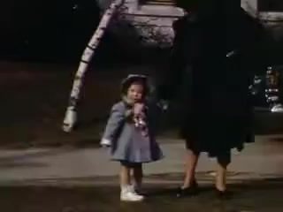 The still from 'Connie and Shirley reel #1 - 1941' features a young girl dressed in a light blue coat and white shoes, holding a toy or snack. She stands next to a woman in a black dress, suggesting a familial or caring relationship. The setting appears to be outdoors, possibly in a residential area, with hints of vintage architecture in the background. The colors are vibrant, characteristic of Kodachrome film from that era.