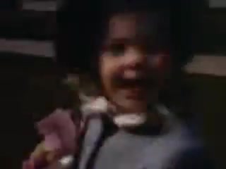 The still from 'Connie and Shirley reel #1 - 1941' features a small child joyfully smiling, appearing to be outdoors. She is dressed in light clothing with a playful expression, likely holding a toy. The color is vibrant, characteristic of Kodachrome film from that era, capturing a warm and nostalgic moment. The image has a soft focus, common in older film techniques.