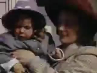 The still features a woman holding a young child, both adorned in large hats. The woman is dressed in a patterned outfit, and the child is in a gray coat, reflecting the style of the early 1940s. The colors are vibrant, characteristic of Kodachrome film, capturing a moment of familial interaction. The background appears to be undefined, emphasizing the connection between the two subjects.