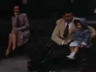 The still captures a moment from 1941, featuring a man dressed in a light-colored suit sitting next to a young girl in a blue outfit. They appear to be sharing a relaxed moment, while a woman in a gray coat is seated nearby, smiling. The vibrant colors typical of Kodachrome film give the scene a nostalgic warmth, suggesting a casual family outing or gathering.